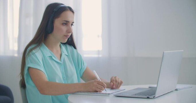 student woman sit at desk at home wear headset talk distantly by video call app and laptop device e-learn language studying with e tutor or classmate do common task remotely, modern tech concept