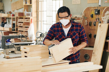 Mature Asian carpenter in goggles and medical mask sanding wooden crates he made for customer