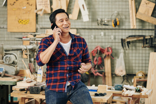 Cheerful mature Asian carpentry shop owner talking on phone with client