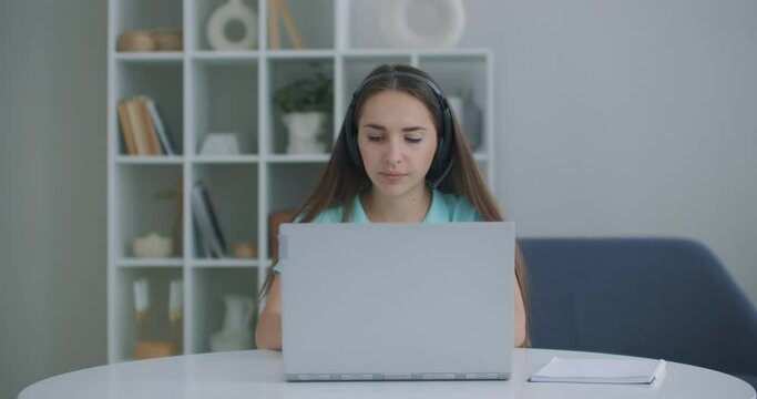 student woman sit at desk at home wear headset talk distantly by video call app and laptop device e-learn language studying with e tutor or classmate do common task remotely, modern tech concept