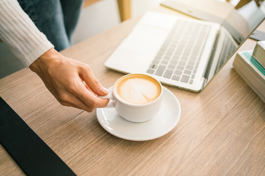 Crop image of a woman hand wearing a sweater holding a morning cup of hot latte coffee, laptop and novels on a wooden working table. Work from home concept. Quarantine period.