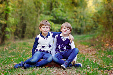 Fototapeta premium Portrait of little school kids boys sitting in forest. Happy children, best friends and siblings having fun on warm sunny day early autumn. Twins and family, nature and active leisure.