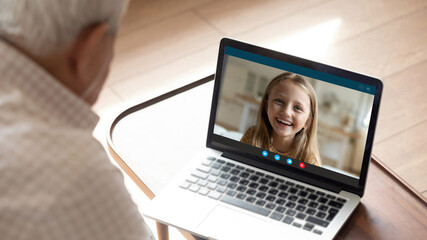 Close up rear view of elderly grandfather speak talk on video call on laptop with smiling small granddaughter. Mature grandparent have webcam digital conference with happy grandchild on computer.