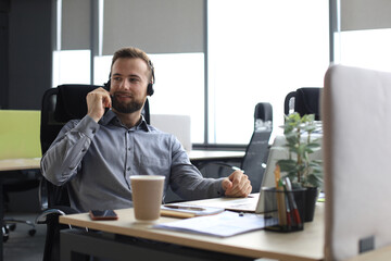 Smiling male call-center operator with headphones sitting at modern office, consulting online...