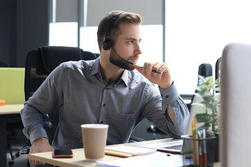 Smiling male call-center operator with headphones sitting at modern office, consulting online...