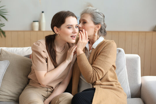 Happy middle mother whispering secret to her smiling daughter at home, gossiping, sharing secret. Beautiful woman hearing good news from mature mum, trusted relations.