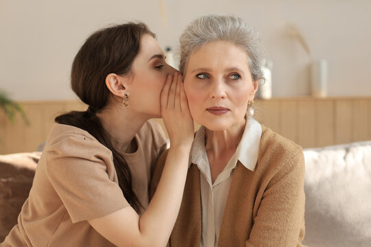 Happy adult daughter whispering secret to her middle mother at home, gossiping, sharing secret. Beautiful woman hearing good news from mature mum, trusted relations.
