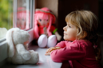 Fototapeta na wymiar Cute toddler girl sitting by window and looking out on rainy day. Dreaming child with doll and soft toy feeling happy. Self isolation concept during corona virus pandemic time. Lonely kid.