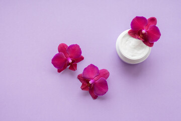 White container with cream for face and body with three magenta colored orchid flowers on purple background. Concept of delicate or eco friendly cosmetics