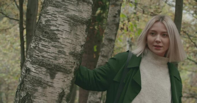 young shy blonde woman walks on the leafs around a tree in autumn. Slow motion