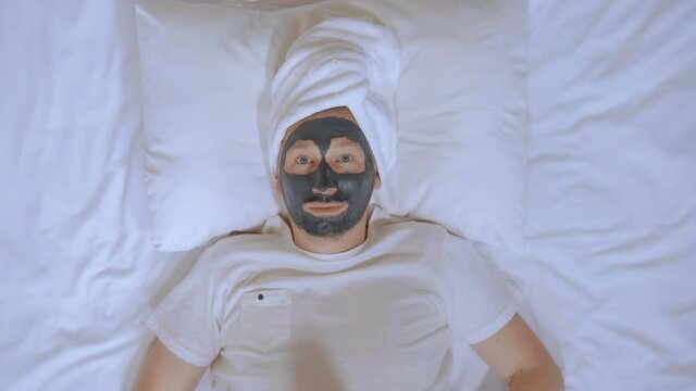 top view adult caucasian man with black mudpack holding paper smiley face in bedroom looking at the camera happy smiling spa treatments at home