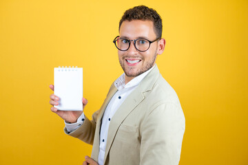 Young handsome businessman wearing suit over isolated yellow background smiling and showing blank notebook in his hand