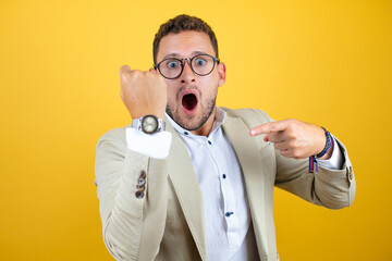 Young handsome businessman wearing suit over isolated yellow background surprised and pointing his...