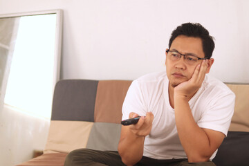 Bored tired  young Asian man sitting on the sofa and watching TV. Trying to choose interesting TV channel