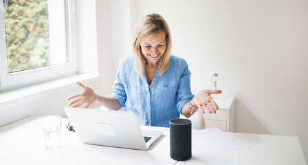 beautiful young and blond woman is talking to her smart speaker and is working at home during...