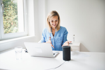 beautiful young and blond woman is talking to her smart speaker and is working at home during...