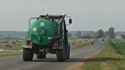 Old wheeled tractor with green sewage disposal barrel on trailer move on asphalted rural road on field at autun day, farming mechanization machines transport equipment, back view