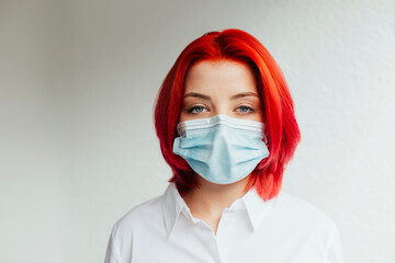 Portrait of a cool young girl with bright red hair and surgical face mask because of coronavirus looking at camera