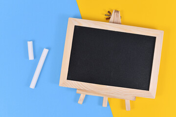 Empty wooden black chalkboard next to pieces of white chalk on yellow and blue background