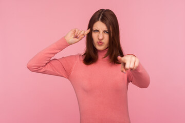 Obraz na płótnie Canvas You are stupid! Sad dissatisfied brunette woman showing stupid gesture pointing finger at you looking at camera, blaming and mocking. Indoor studio shot isolated on pink background