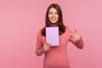 Fototapeta na wymiar Positive smiling woman pointing finger showing notebook with blank space for commercial text, advertising area on paper for plans. Indoor studio shot isolated on pink background