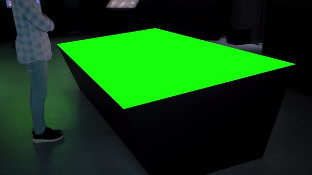 Woman looking at blank digital interactive green display of table kiosk at exhibition or museum with futuristic sci-fi interior. Mock up, copyspace, template, chroma key, green screen concept