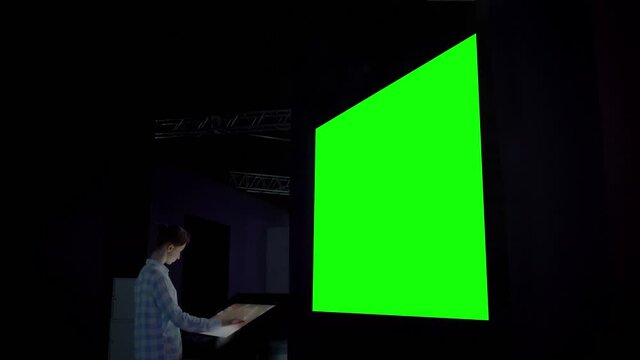 Green screen, mock up, futuristic, chroma key, template concept. Woman using electronic kiosk and looking at green blank large interactive wall display in dark room of modern technology museum
