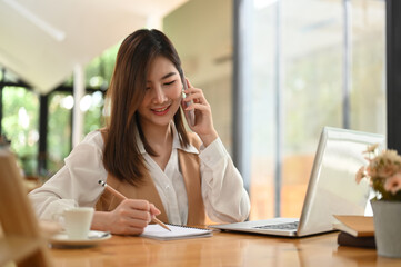 Young female talking on the phone while writing on notebook at office.