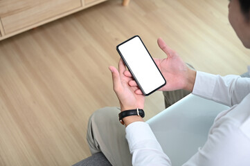 Young male freelancer holding phone while looking at screen watching social media on sofa.