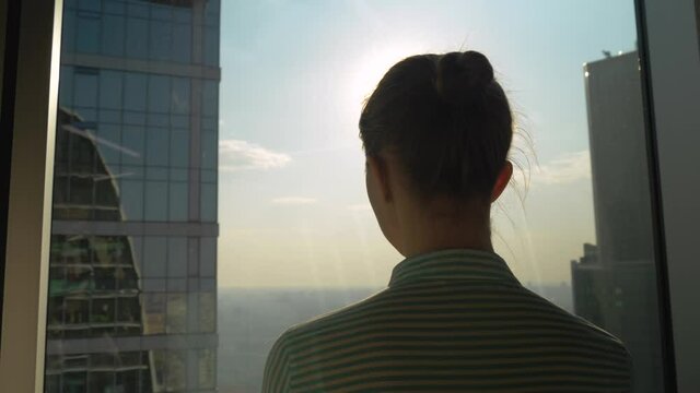 Back view of woman silhouette looking at cityscape through window of skyscraper. Summer time, sunset, sun lens flare. Success, opportunity, sightseeing, discover and future concept