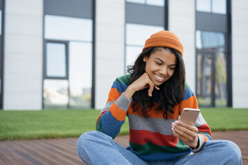 Beautiful smiling woman using mobile phone, internet, watching movie online, sitting in park. Happy African American student studying, learning languages, online education concept