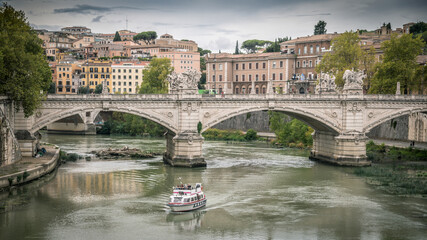 Fototapeta premium Panoramic view Emanuele II bridge over Tevere river in Rome, near Castel Sant'Angelo. Boat sails on the river of the Italian capital, historic buildings in the background. Tourism, art concept