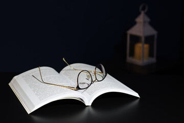 Golden glasses on open book by dark background with night lantern