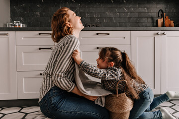 Daughter indulges and tickles mom. Woman and child laugh and pose on kitchen floor with laundry...