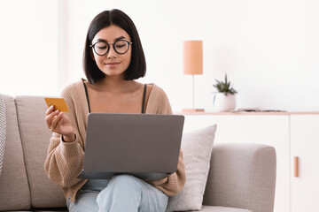 Asian woman making purchases sitting with pc on couch