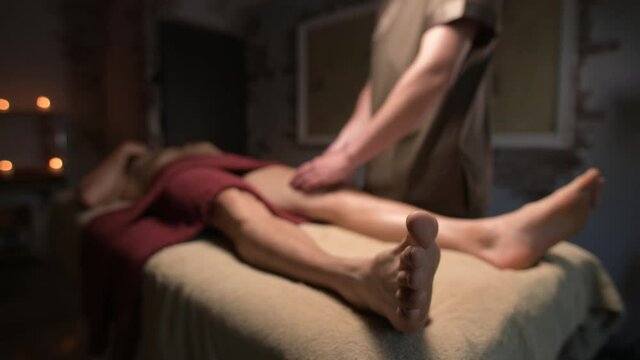 Male masseur doing sports massage of the thighs of the legs to a muscular athlete in an office with a dim light
