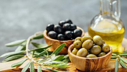 Set of green and black olives and olive oil on gray background. Different types of olives in olive wooden bowls and olive oil over wooden cutting board and olive leaves. Copy space. Horizontal banner