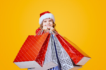 Adorable child in red Santa hat and sweater with deer carrying colorful shopping bags with Christmas gifts on yellow background.