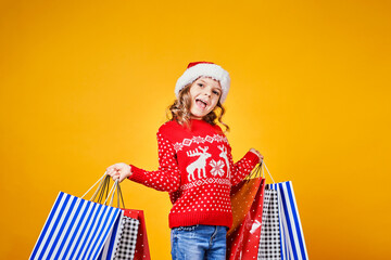 Adorable child in red Santa hat and sweater with deer carrying colorful shopping bags with Christmas gifts on yellow background.