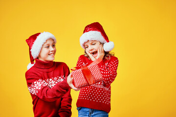 Happy boy in Santa hat giving Christmas gift box to excited girl in red sweater with deer on yellow background