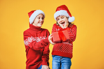 Happy boy in Santa hat giving Christmas gift box to excited girl in red sweater with deer on yellow background