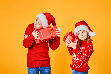 Cheerful kids in Santa hats and red sweaters standing with presents laughing and looking at camera on yellow background