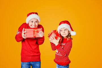 Cheerful kids in Santa hats and red sweaters standing with presents laughing and looking at camera on yellow background