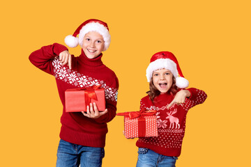 Cheerful kids in Santa hats and red sweaters standing with presents laughing and looking at camera on yellow background
