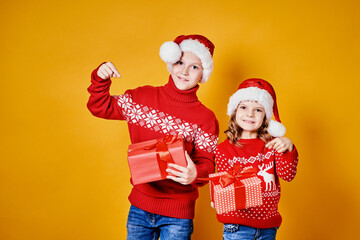 Cheerful kids in Santa hats and red sweaters standing with presents laughing and looking at camera on yellow background
