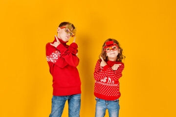 Children in warm red Christmas sweaters and decorated glasses looking at camera on yellow background.