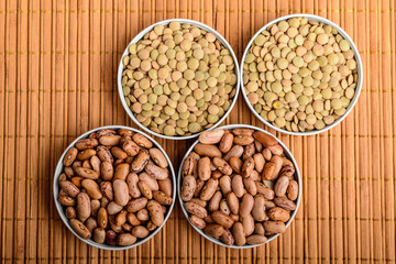 Top view of four round bowls with many fresh organic red beans and green lentils on a wooden table in warm light, brown monochrome indoor background photographed with selective focus.