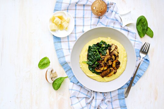 A portion of polenta or corn grits with fried mushrooms and stewed spinach in a gray plate on a light concrete background. Polenta or corn grits recipes.