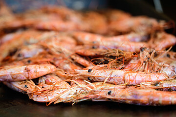 Close up of large portion of cooked shrimps on black grill at a street food festival, ready to eat healthy seafood, beautiful orange monochrome outdoor background.