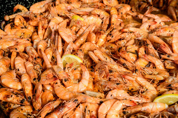 Close up of large portion of cooked shrimps with tomato sauce and yellow lemons in a large pan at a street food festival, ready to eat healthy seafood, beautiful orange monochrome outdoor background.